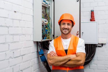 Handsome electrician with crossed arms smiling at camera near electric panel clipart