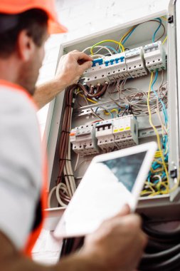 Selective focus of electrician in hardhat and safety vest holding digital tablet and fixing electric panel clipart