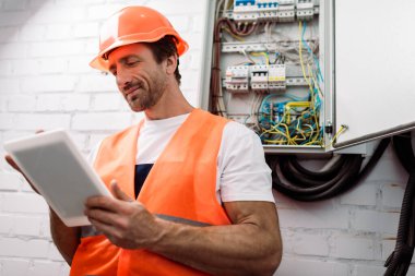 Selective focus of handsome electrician using digital tablet near electrical distribution box clipart