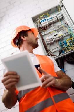 Selective focus of handsome electrician in hardhat looking at electrical distribution box while holding digital tablet  clipart