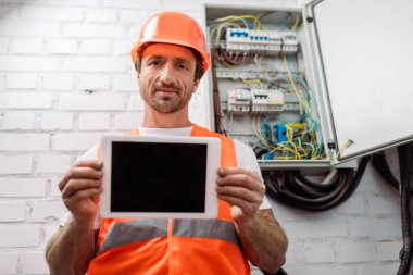 Selective focus of handsome electrician showing digital tablet with blank screen near electrical box clipart