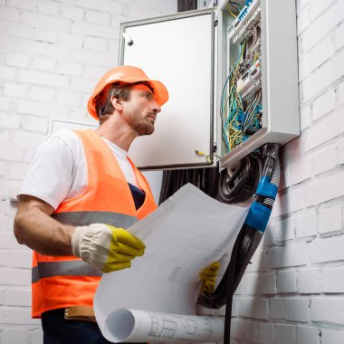 Şablonu tutan yakışıklı elektrikçi ve elektrik dağıtım kutusuna bakıyor.