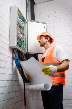 Kask ve güvenlik yeleği giymiş bir elektrikçi elinde planlar ve elektrik paneline bakıyor.