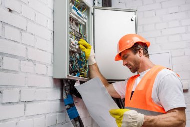 Şablona bakıp elektrik dağıtım kutusunu tamir eden eldivenli yakışıklı bir elektrikçi.