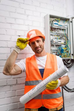 Handsome repairman in hardhat looking at camera while holding blueprint near electrical distribution box clipart