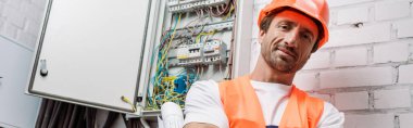 Panoramic shot of electrician with blueprint looking at camera near electrical box clipart