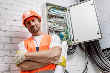 Handsome workman in helmet and safety vest holding blueprint and looking at camera near electric panel clipart
