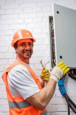 Elektrik dağıtım kutusunun yanında kabloları tutarken kameraya gülümseyen kasklı elektrikçinin yan görüntüsü