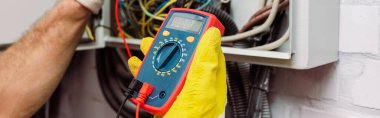 Panoramic crop of electrician using multimeter near electrical distribution box clipart