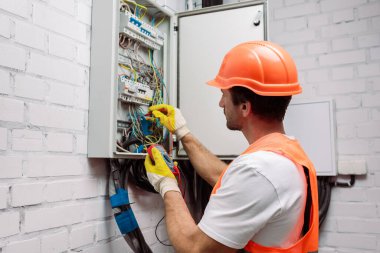 Handsome electrician in hardhat and gloves holding multimeter near electrical distribution box clipart
