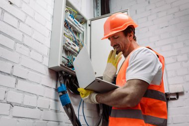Elektrikli panelin yanında dizüstü bilgisayar tutarken eldivenini çıkaran yakışıklı elektrikçinin yan görüntüsü