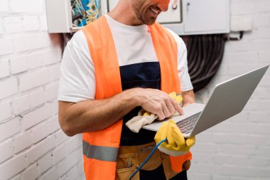 Cropped view of smiling electrician holding glove and using laptop  clipart