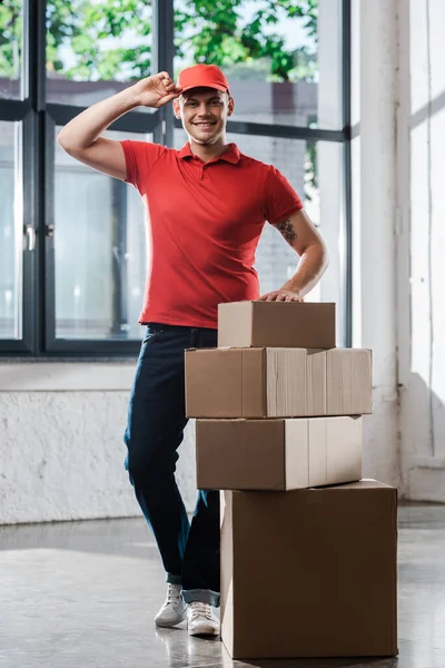 Happy Tattooed Delivery Man Touching Cap While Standing Carton Boxes — Stock Photo, Image