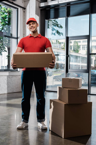 cheerful delivery man in cap holding carton box and looking at camera