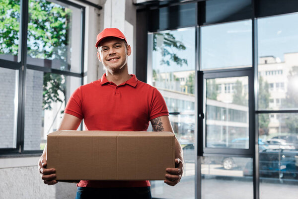 positive delivery man in cap holding carton box and looking at camera