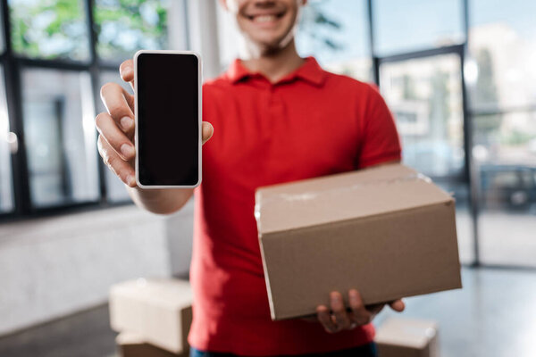 cropped view of smiling delivery man holding smartphone with blank screen and carton box 