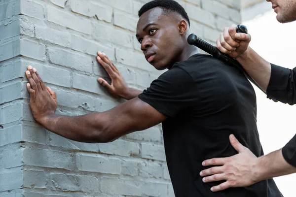 african american man standing against brick wall near policeman with truncheon, racism concept