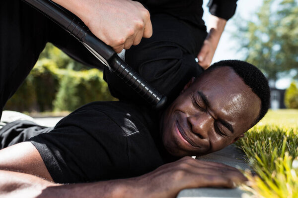 selective focus of detained african american man suffering from pain while lying near policeman with baton, racism concept 