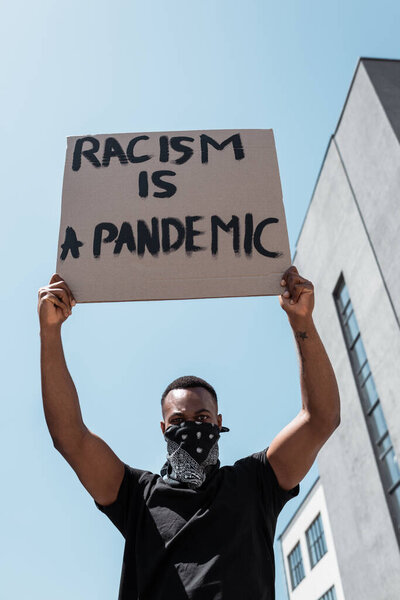 low angle view of african american man with scarf on face holding placard with racism is a pandemic lettering on street