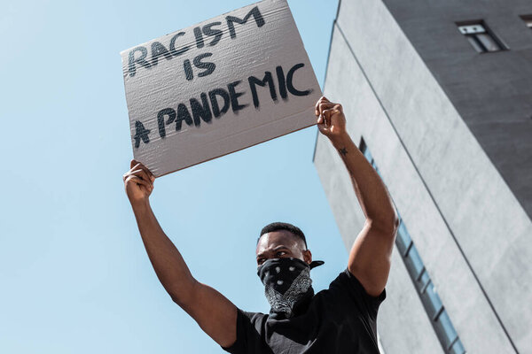 low angle view of african american man with scarf on face holding placard with racism is a pandemic lettering near building 