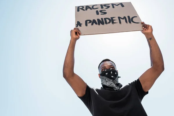 非洲裔美国人脸上戴着围巾 手持种族主义标语牌的低视角形象 是一种在蓝天映衬下的大流行病 — 图库照片