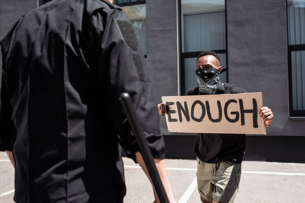 selective focus of african american man with scarf on face holding placard with enough lettering near policeman with baton on street, racism concept 