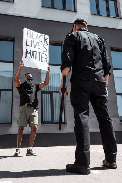 selective focus of african american man with scarf on face holding placard with black lives matter lettering near policeman with baton on street, racism concept 