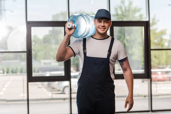 cheerful delivery man in cap and uniform holding gallon of water in bottle