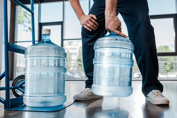 cropped view of delivery man holding heavy bottle with water near hand truck