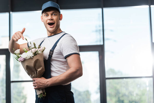 surprised delivery man in uniform and cap pointing with finger at bouquet with flowers 