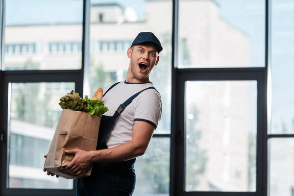 excited delivery man in cap holding paper bag with groceries 