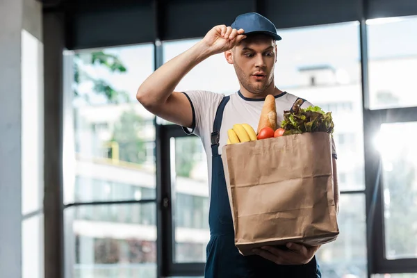 Sürpriz Teslimatçı Şapkaya Dokunuyor Poşetteki Yiyeceklere Bakıyor — Stok fotoğraf