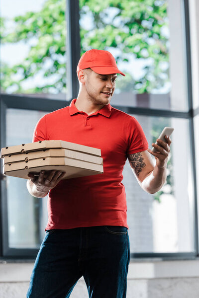 delivery man in cap looking at smartphone and holding pizza boxes 