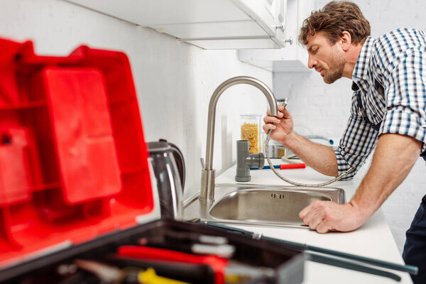 Selective focus of plumber holding metal pipe near kitchen faucet and toolbox 