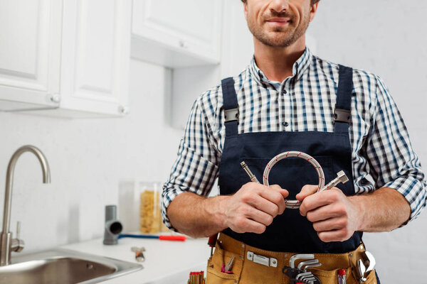 Cropped view of plumber in workwear and tool belt holding metal pipe in kitchen 