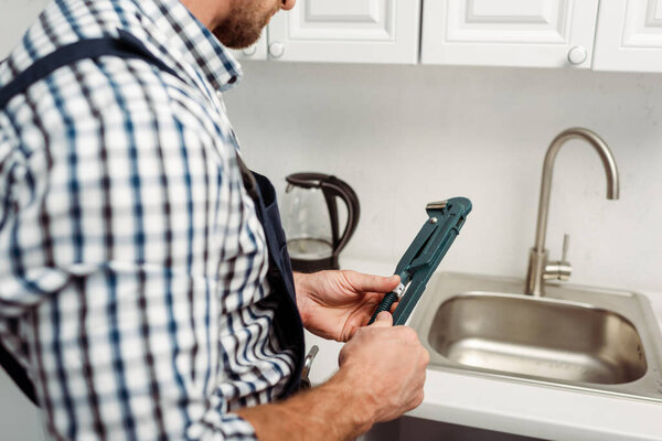 Cropped view of plumber holding wrench near faucet and sink in kitchen 