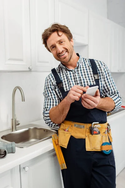 Lachende Loodgieter Met Behulp Van Smartphone Buurt Kraan Wastafel Keuken — Stockfoto