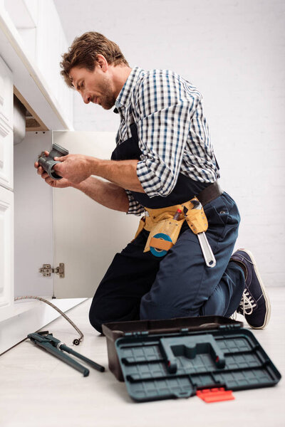 Selective focus of handsome plumber holding plastic pipe near pipe wrench and toolbox on floor in kitchen 