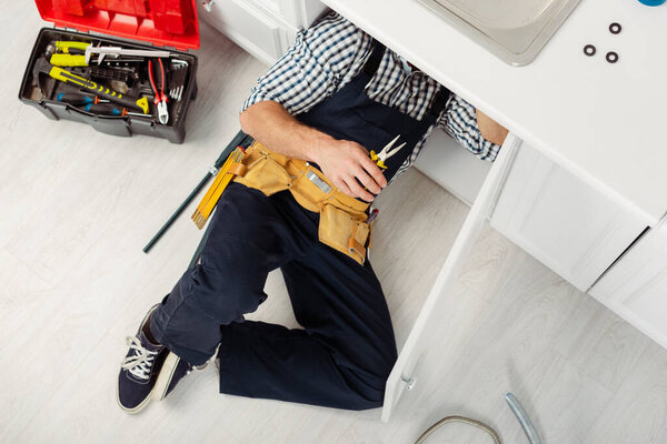 Top view of handyman in overalls and tool belt holding pliers while repairing sink in kitchen 