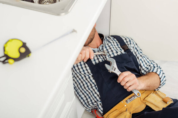 Selective focus of plumber in overalls holding wrench and metal pipe while fixing kitchen sink 