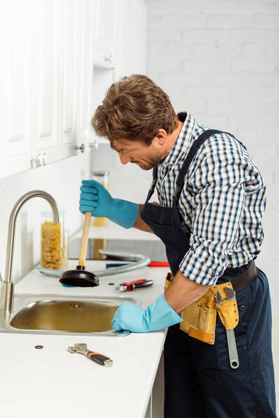 Side view of plumber in rubber gloves holding plunger while fixing kitchen sink 