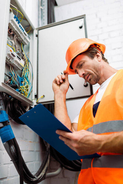 Selective focus of handsome electrician in hardhat holding pen and looking at clipboard near electric panel
