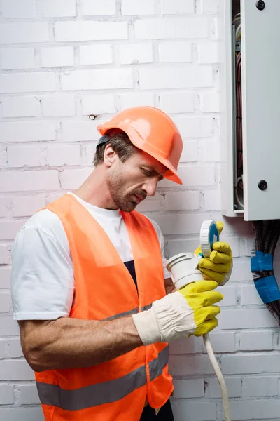 Handsome electrician in gloves repairing wiring near electrical distribution box