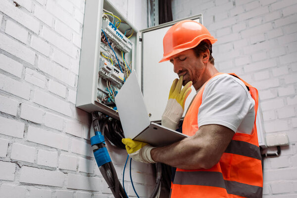 Side view of handsome electrician taking off glove while holding laptop near electric panel