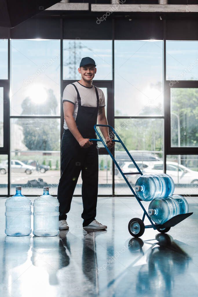 cheerful delivery man in uniform holding hand truck with bottled water 