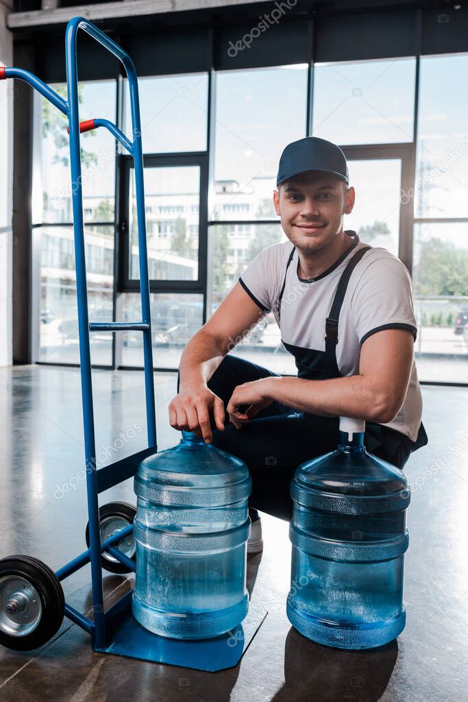 happy delivery man in uniform sitting near hand truck with purified water in bottles 