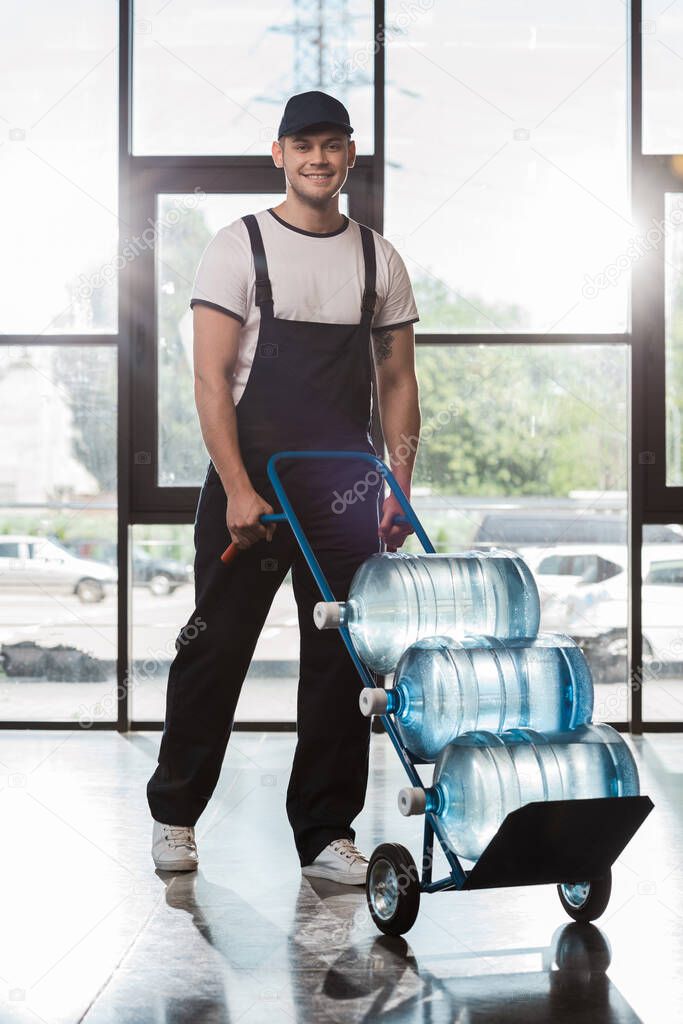 cheerful delivery man in cap and uniform holding hand truck with bottled water 