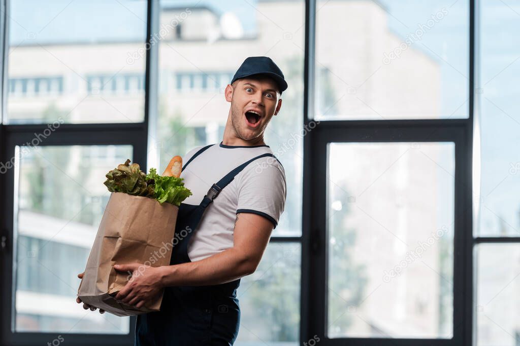 excited delivery man in cap holding paper bag with groceries 