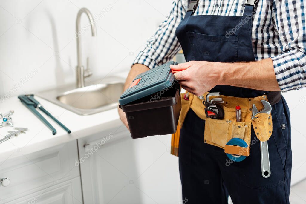 Cropped view of plumber with tools on tool belt holding toolbox in kitchen 