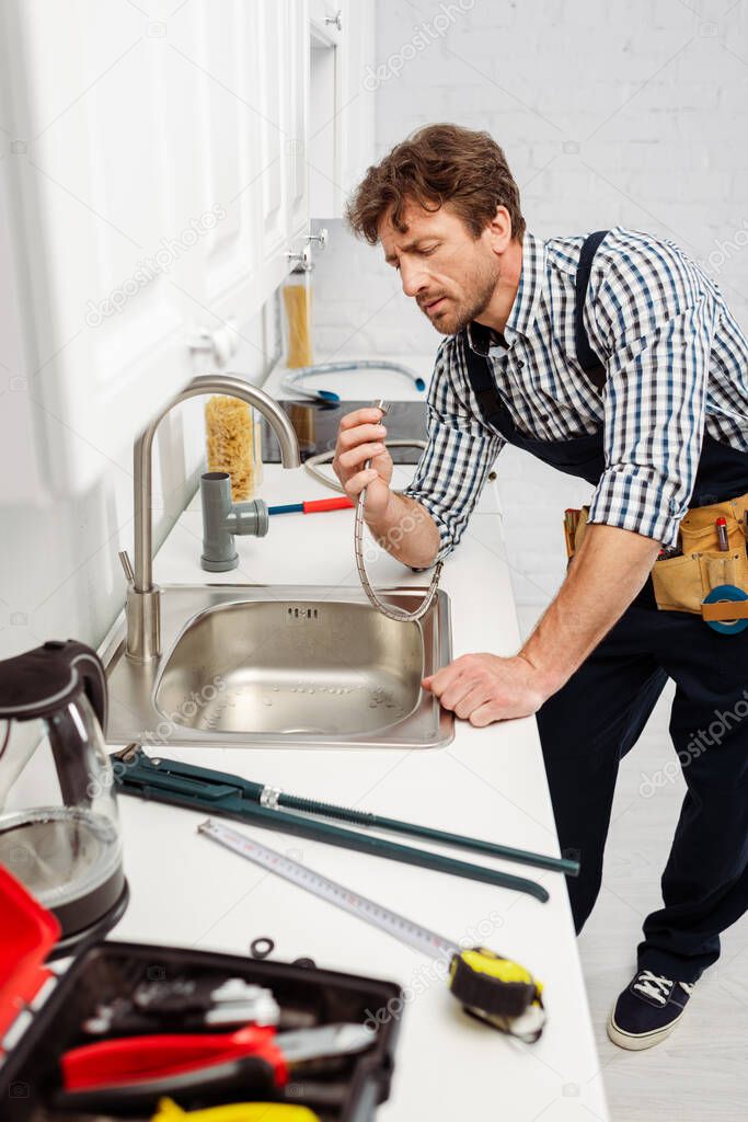 Selective focus of plumber in overalls holding metal pipe near sink in kitchen 